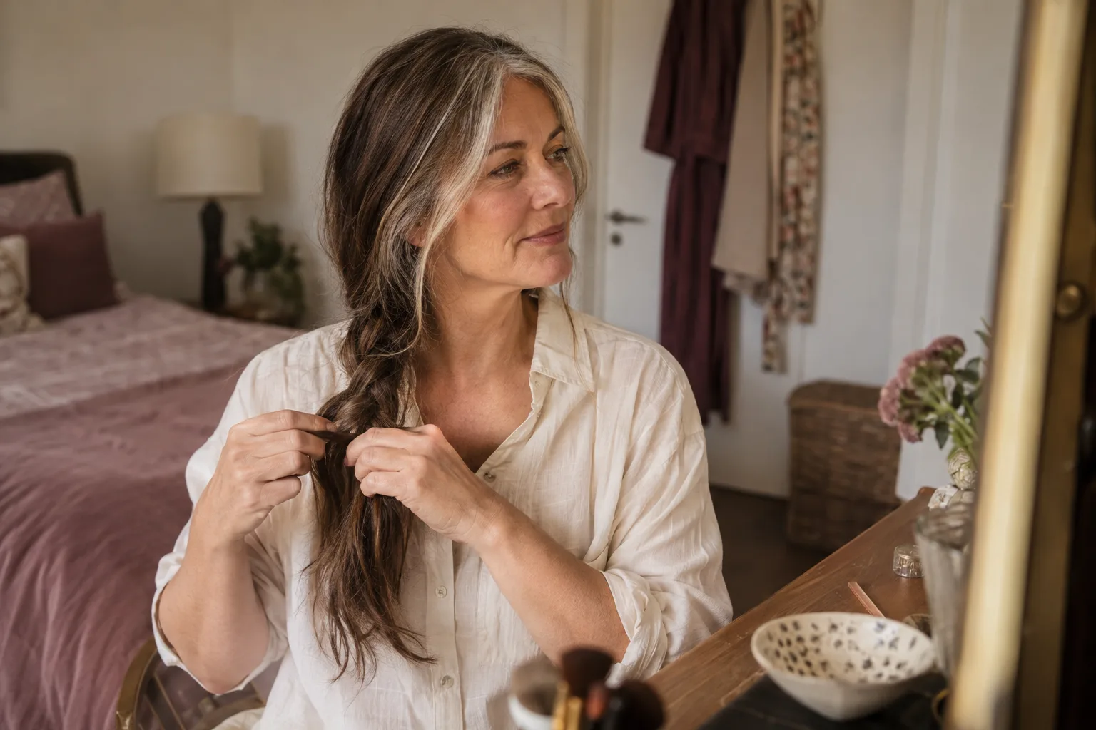 Woman with long grey hair and brunette lengths forming a loose side plait beside a bedroom dressing table